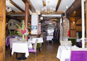 A restaurant with wooden beams, a brick wall, and a chandelier. Tables are set with white tablecloths and purple chairs. - Hôtel du Mont Blanc (Paris)