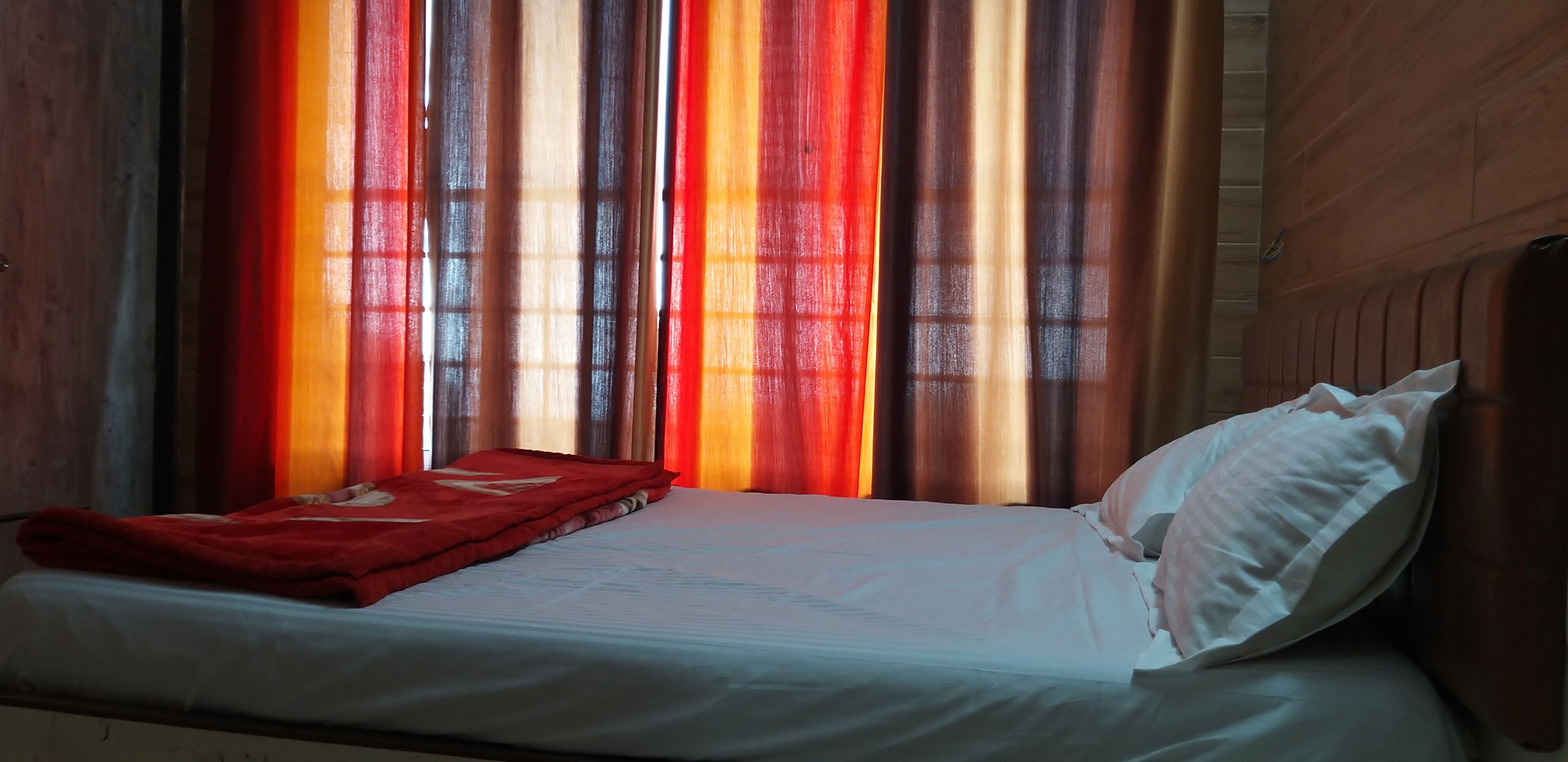 Ein Bett mit weißer Bettdecke und roter Decke, ein Holz-Kopfbrett und Vorhänge mit vertikalen Streifen.
