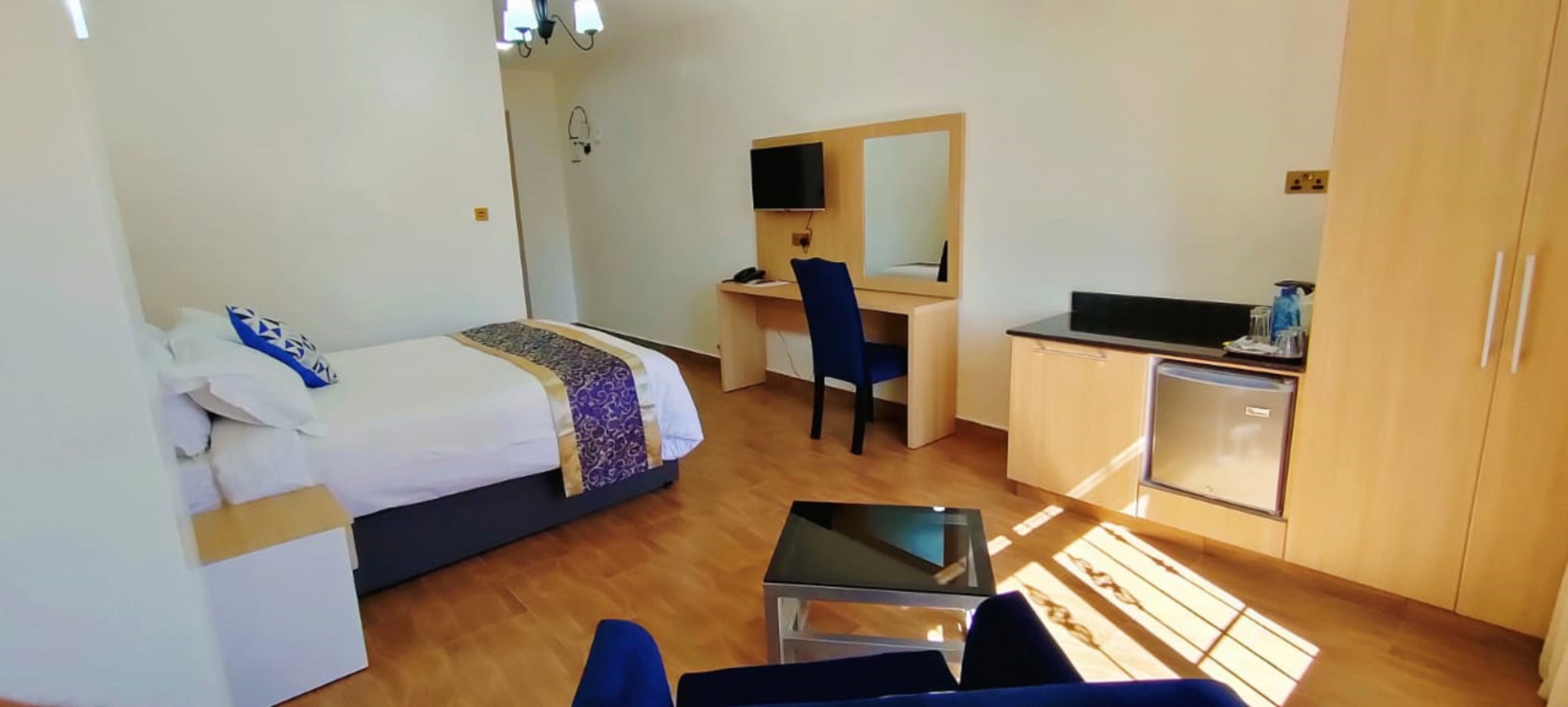 A hotel room with a bed, a desk with a chair, a small kitchenette, and a TV mounted on the wall.