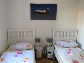 Une chambre avec deux lits, chacun agrémenté de draps à motifs floraux, une table de chevet avec une lampe, une petite table et un tableau représentant un phoque, accroché au mur.