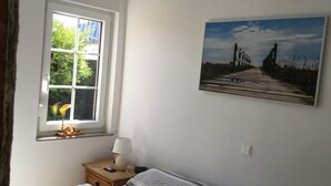 Un dormitorio con ventana, una mesita de noche con lámpara, una cama y un cuadro colgado en la pared.
