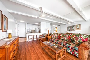 A spacious living area with a wooden floor, a colorful sofa, a glass coffee table, and a dining area in the background.