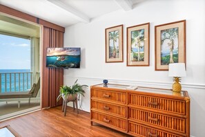 A room with a wooden floor, a large window overlooking the sea, a television mounted on the wall, and a wooden cabinet with drawers.
