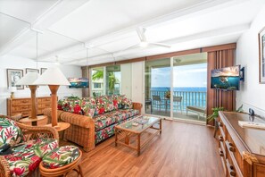 Un'ampia zona giorno con divano, tavolino da caffè e mobili in vimini. Una televisione è montata a parete, e la stanza dispone di un'ampia porta scorrevole in vetro che dà su un balcone con vista sull'oceano.