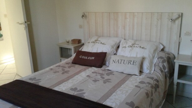 Un lit recouvert d’une couvre-lit à motifs floraux, deux oreillers ornés du mot « NATURE » et un autre du mot « COULEUR », une petite table de chevet et une commode.