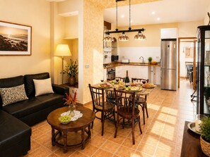A living room with a black sofa, a dining table with chairs, a kitchen area with a refrigerator, and a dining table set with food and drinks.