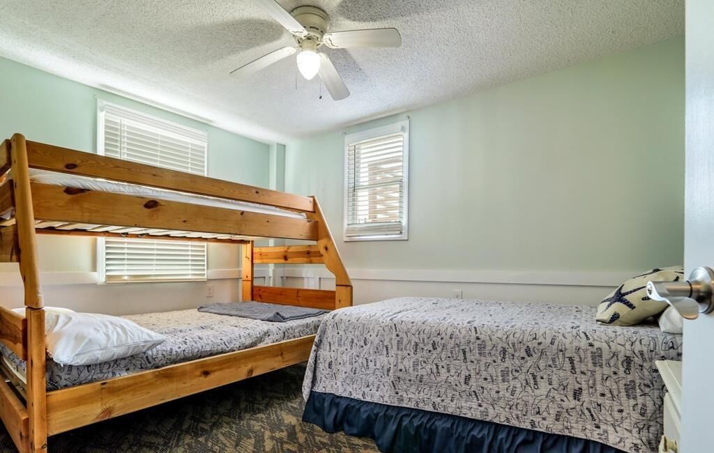A room with two bunk beds, a ceiling fan, and two windows with blinds.