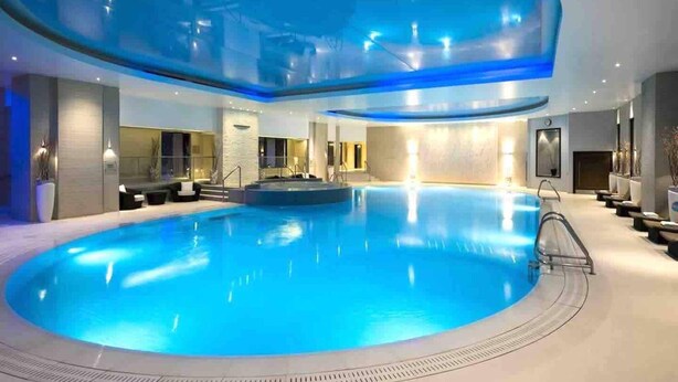 A large indoor swimming pool with a curved ceiling, surrounded by seating areas and a reception desk.
