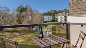 A balcony with a wooden table and chairs, a bottle of wine, and a glass. In the background, there are trees and a building with multiple windows.