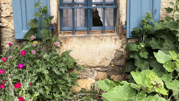 Eine rustikale Steinmauer mit einem kleinen Fenster, ausgestattet mit blauen Fensterläden und einem Vorhang.