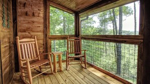 Uma cabana de madeira com duas cadeiras de balanço e uma mesa pequena, com vista para uma floresta.