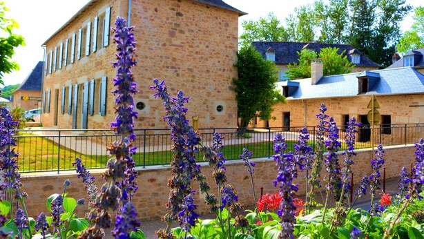 A stone building with a prominent tower and a fenced garden with purple flowers. - Gîte des 3 pays, ideal for reunions with family or friends (cublac)