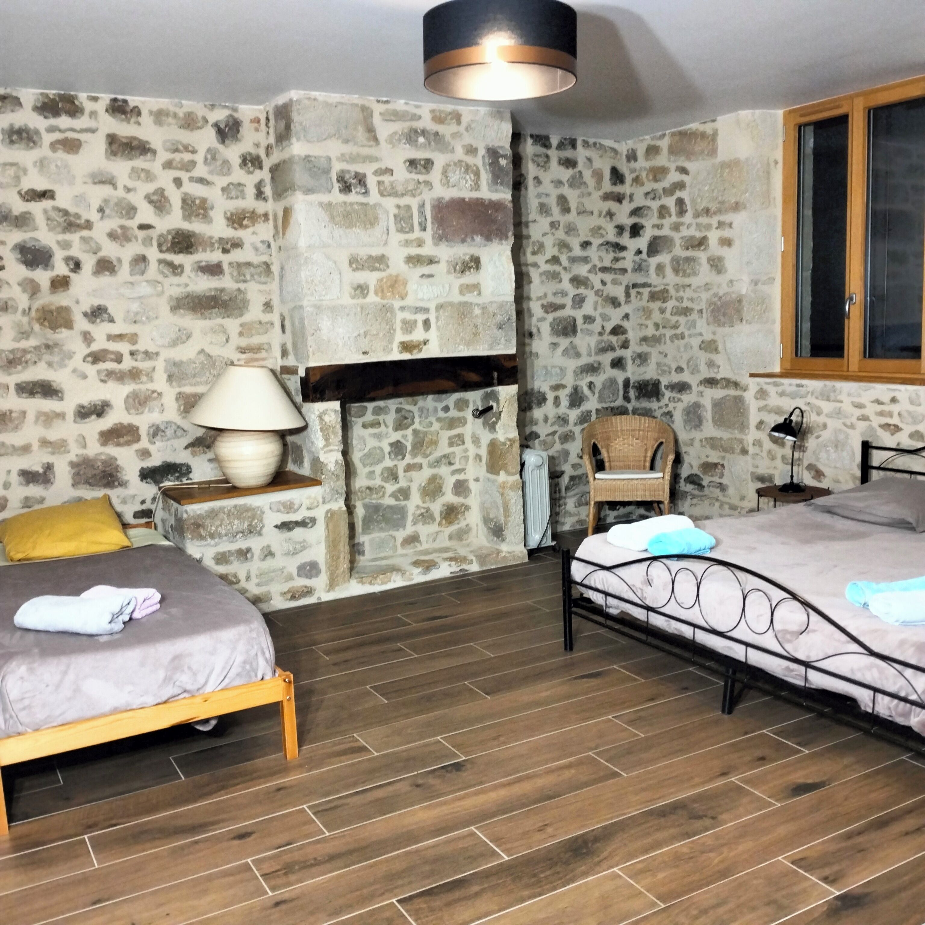 Une chambre avec un mur en pierre, une cheminée, deux lits, une chaise et une fenêtre.