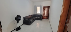 A room with a black leather sofa, a standing fan, a wooden door, and a window with curtains.