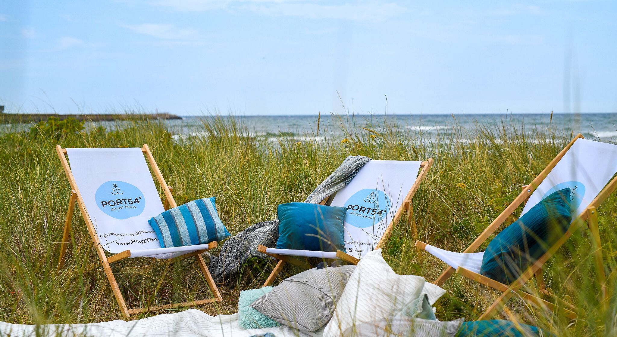 Drie strandstoelen met 'PORT54'-logo, een deken en kussens op een gazon aan de zee.