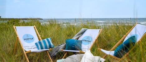 Drie strandstoelen met 'PORT54'-logo, een deken en kussens op een gazon aan de zee.