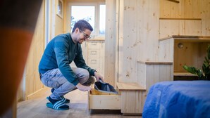 Une personne accroupie dans une pièce en bois, où se trouvent un lit et une petite armoire en bois.