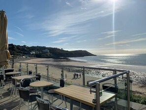 An outdoor seating area with tables and chairs, overlooking a beach with a clear view of the ocean and distant hills.