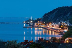 A coastal town at dusk with a pier extending into calm waters, buildings along the shore, and a prominent hill in the background.