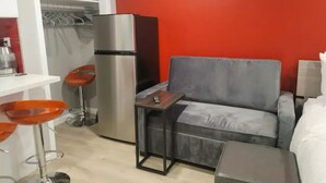 A compact living space with a grey sofa, a small table, a refrigerator, and a kitchen area with a white countertop and orange chairs. - Modern Luxury Oasis Near San Francisco/Oakland (Hayward)