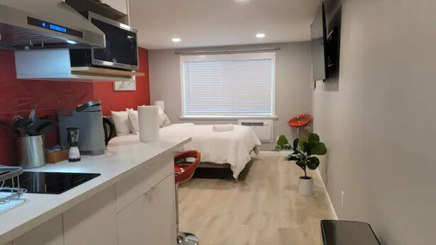 A modern kitchen with a built-in stove, a dining area with a red chair, a bed with white bedding, and a window with blinds. - Modern Luxury Oasis Near San Francisco/Oakland (Hayward)