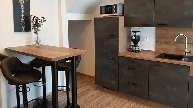 A modern kitchen with dark cabinets, a wooden countertop, a coffee machine, and a microwave. There is a wooden table with two chairs and a vase of flowers on the wall. - Vacation cottage Am Mühlengraben (Merchweiler)