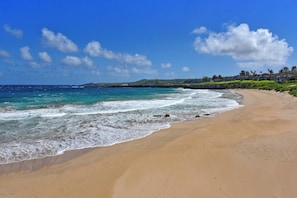 A beach with clear blue water, a sandy shore, and a distant view of houses on a hillside.