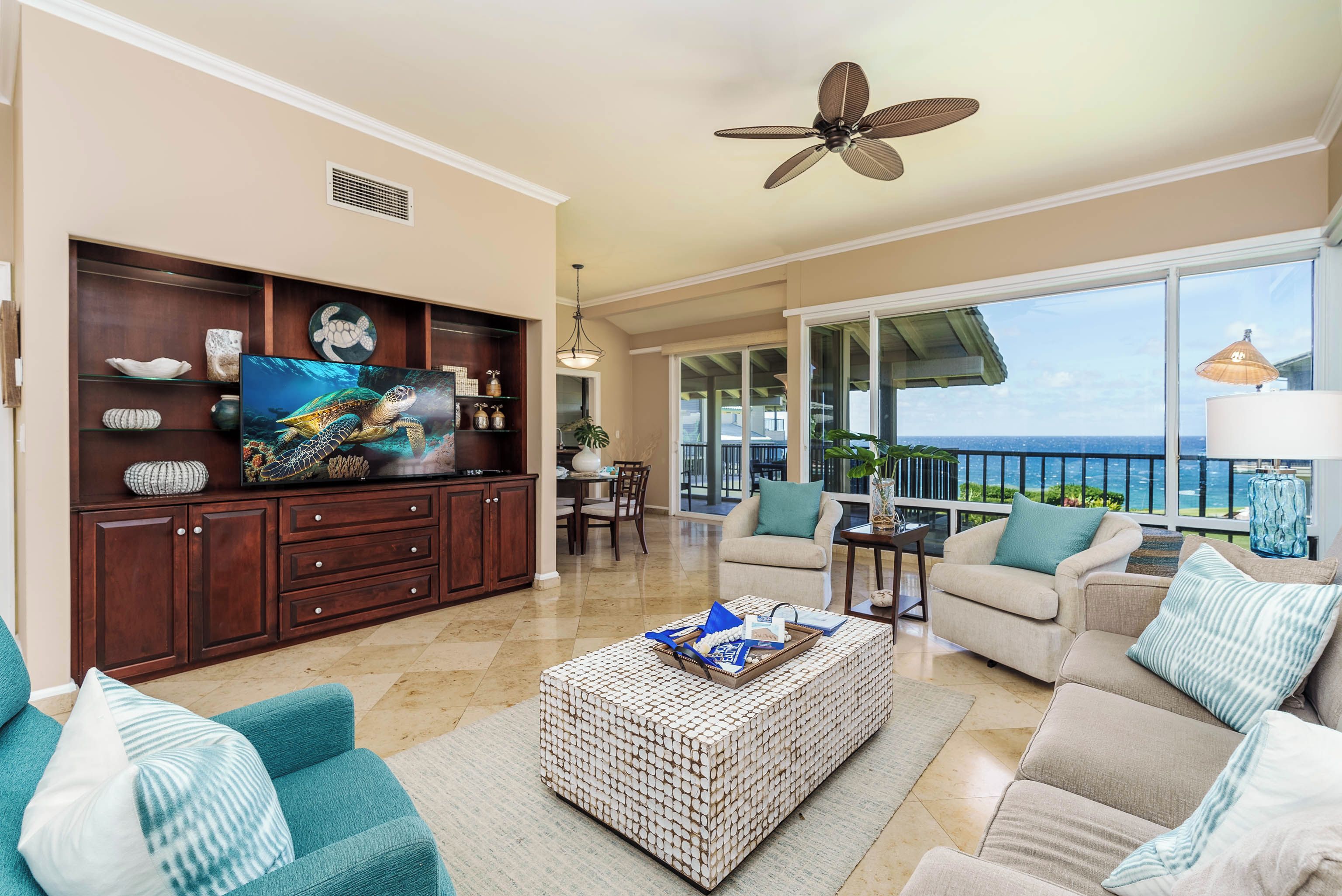 A spacious living room with a large TV, comfortable seating, a ceiling fan, and a view of the ocean.