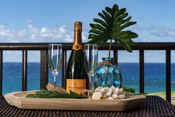 A wooden tray with a bottle of Veuve Clicquot champagne, two glasses, a decorative blue bottle, and a large green leaf against a scenic ocean view.