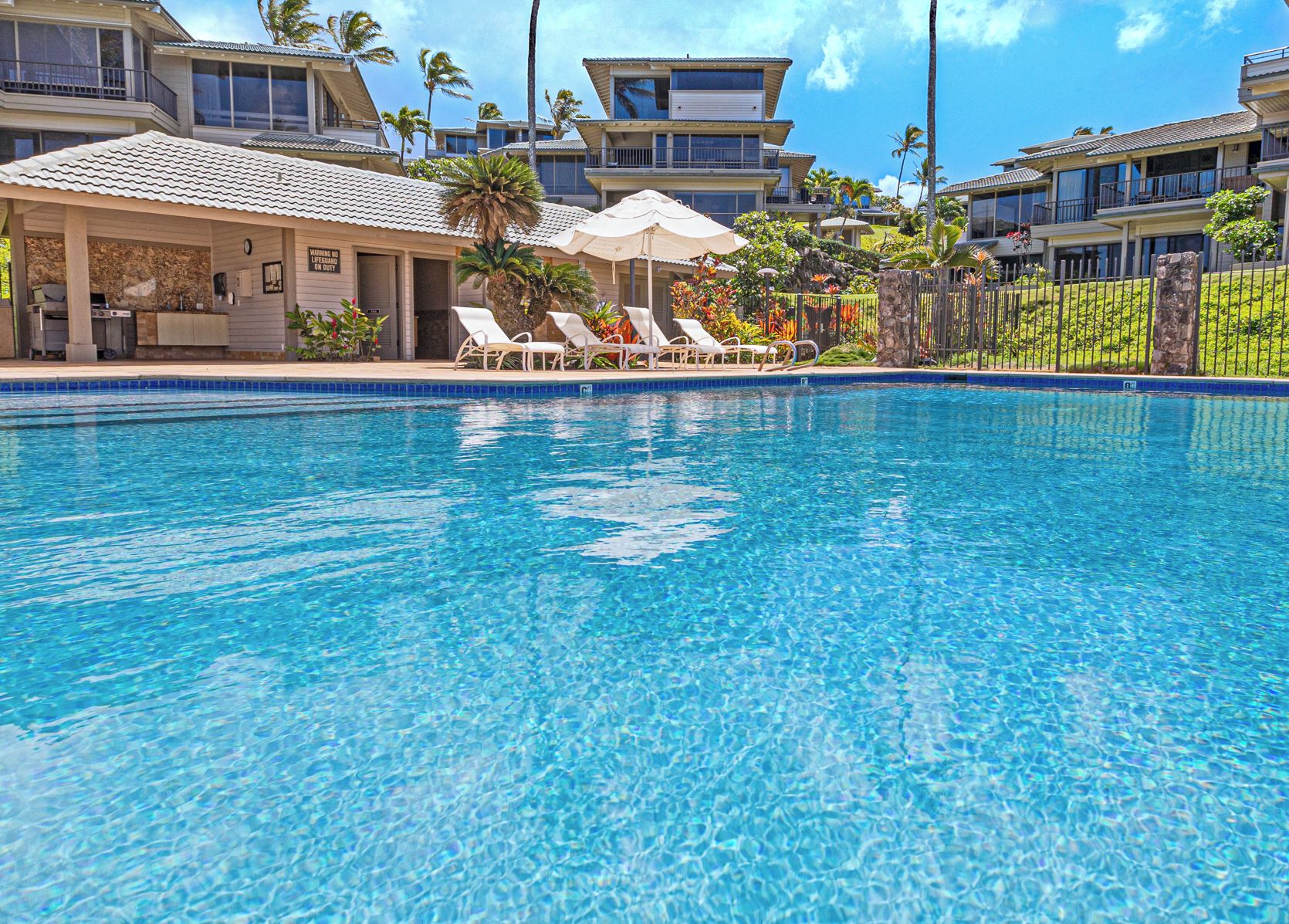 A clear blue pool with lounge chairs and umbrellas, surrounded by multi-story buildings with balconies.