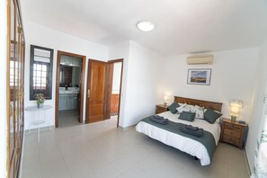 Une chambre à coucher avec un grand lit, des tables de chevet avec des lampes, une armoire en bois, une petite table ronde avec un pot de plante, et une salle de bain visible à travers une porte ouverte.