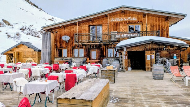 En træchalet med bar og restaurant, udendørs siddepladser med borde og stole, og et sneklædt bjerg i baggrunden.