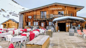 A wooden chalet with a bar and restaurant, outdoor seating with tables and chairs, and a snowy mountain backdrop. - Studio center resort beautiful view Les Coches La Plagne Paradiski (Bellentre)