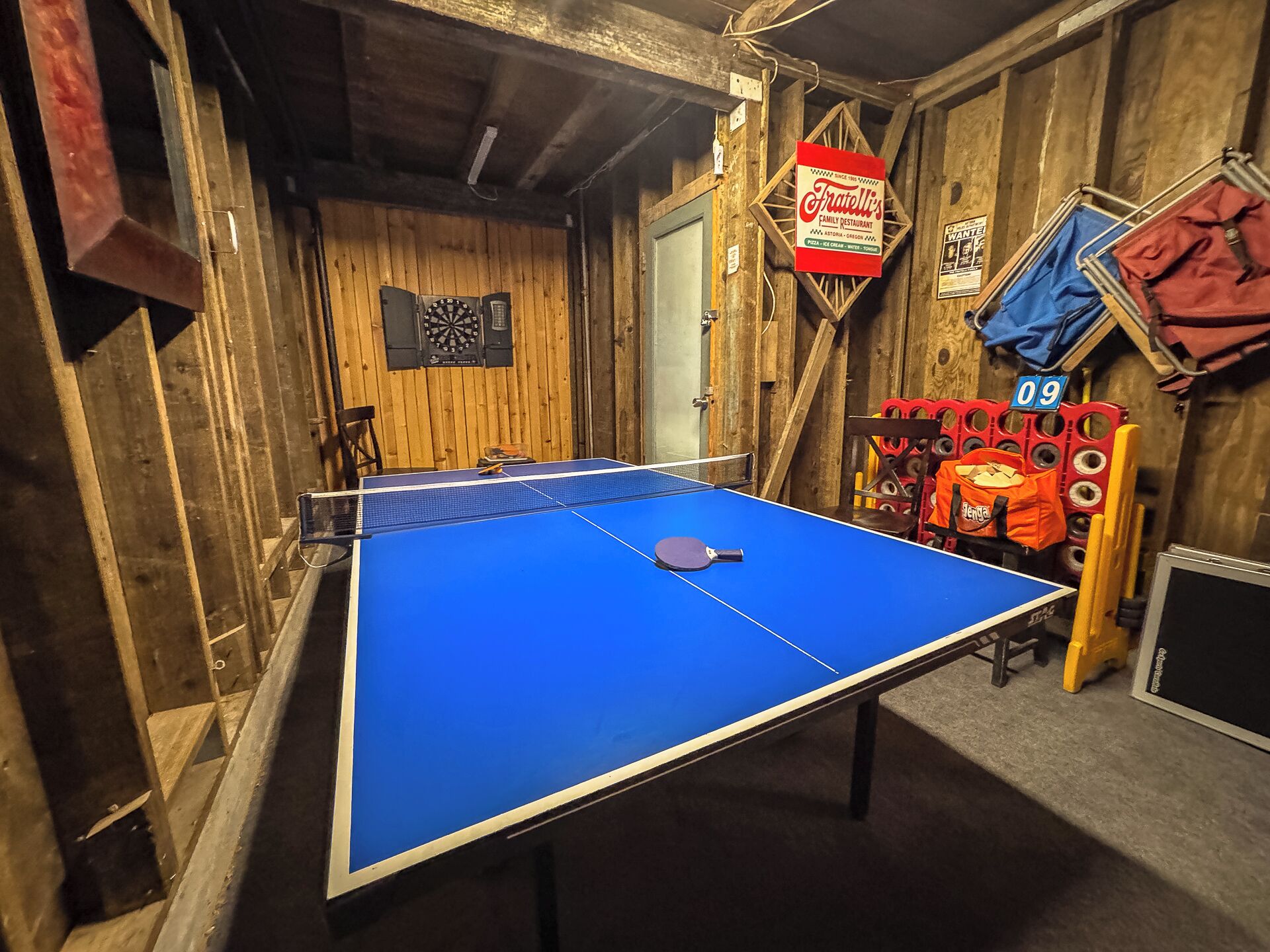Une table de ping-pong dans une pièce rustique aux murs en bois, un jeu de fléchettes et une enseigne indiquant le restaurant familial Fratelli's.