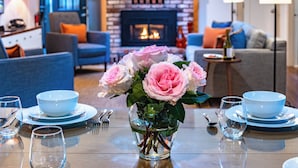 Un'area pranzo con un tavolo apparecchiato per due persone, un vaso di rose rosa e un caminetto sullo sfondo.