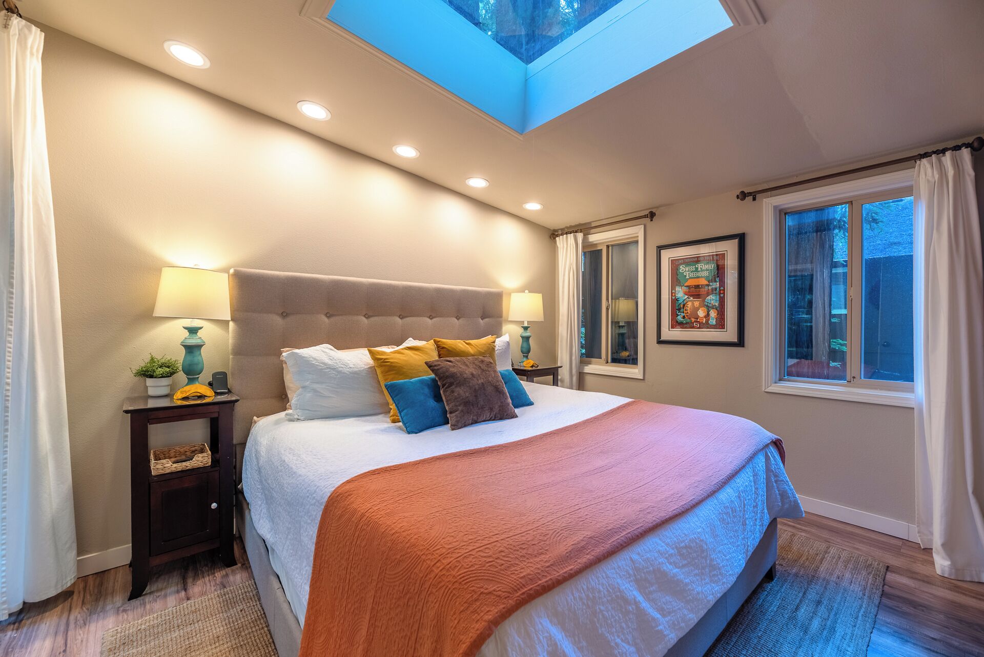 A bedroom with a large bed, two bedside tables, a skylight, a window, and a framed picture on the wall.