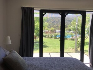 A bedroom with a large bed, a window with curtains, and a view of a pool and greenery outside.