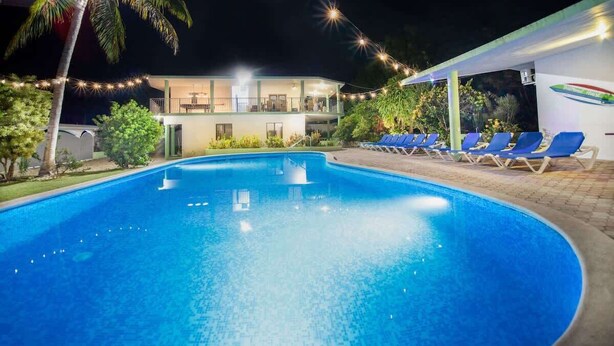 Ein Schwimmbecken mit Liegestühlen, ein Gebäude mit Balkonen und Lichterketten bei Nacht.