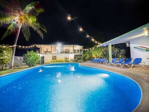Ein Schwimmbecken mit Liegestühlen, ein Gebäude mit Balkonen und Lichterketten bei Nacht.