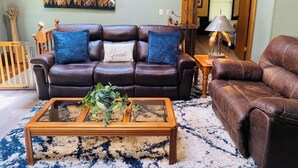 A living room with a brown leather sofa, a glass coffee table with plants, a patterned rug, and a painting on the wall.