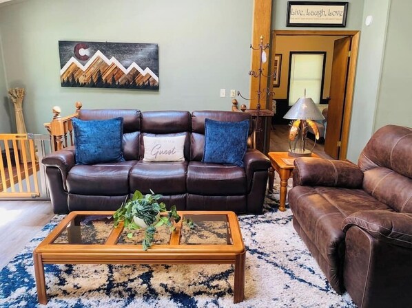 A living room with a brown leather sofa, a glass coffee table with plants, a patterned rug, and a painting on the wall.