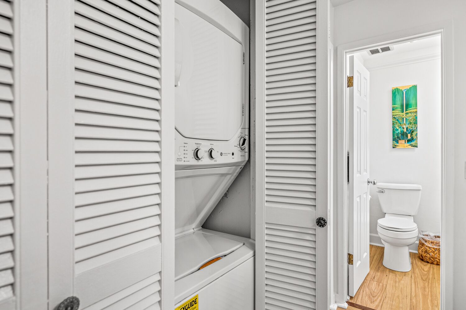 A laundry room with a washing machine, a dryer, and a door leading to a bathroom with a toilet and a painting on the wall.