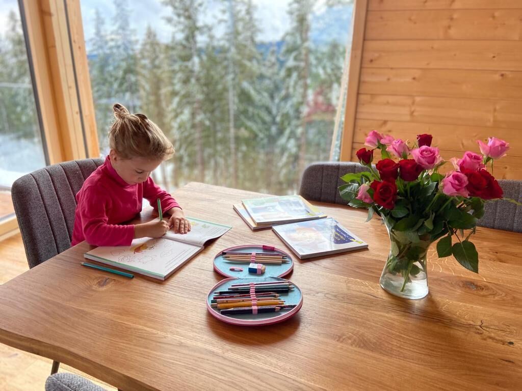 Un niño dibujando en una mesa de madera con vista a la ventana, un jarrón de flores y material de arte.