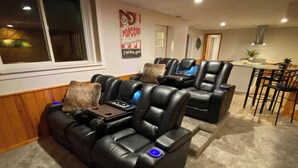 Sistema de cine en casa con dos sillones de cuero reclinables y una mesa de centro, comedor con barra y cocina al fondo.