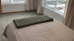 Un dormitorio con una cama grande, ropa de cama color crema y vistas a un paisaje nevado a través de dos ventanas.