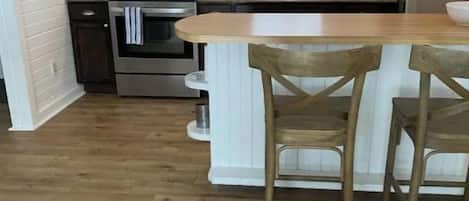 Cocina con paredes de madera blanca, suelo de madera, una isla central con dos sillas y electrodomésticos de acero inoxidable.