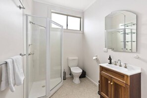 A bathroom with a glass shower enclosure, a toilet, and a vanity with a mirror and shelves. - Barwon Veuve Cottage (Barwon heads)