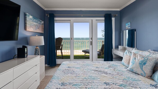A bedroom with a large bed, a TV, a dresser, and a balcony with a view of the ocean.