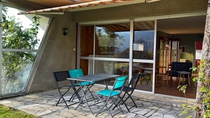 A patio with a table and chairs, a sliding glass door leading to an indoor area with a bookshelf and a chair. - Terrace, Garden, Apartment 80m², mountain view, wifi, office (Bagnères-de-Bigorre)