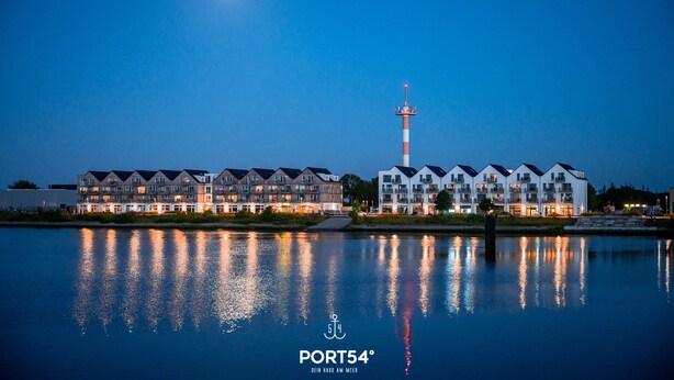 A waterfront hotel with multiple buildings, a prominent tower, and a calm reflective water surface. - Lütte Strandhus - Semi-detached house, fireplace, shower room, washing machine (Olpenitz)